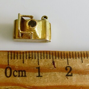 May include: A gold-colored camera charm with a black lens. The charm is about 1 inch long and is sitting on a wooden ruler.