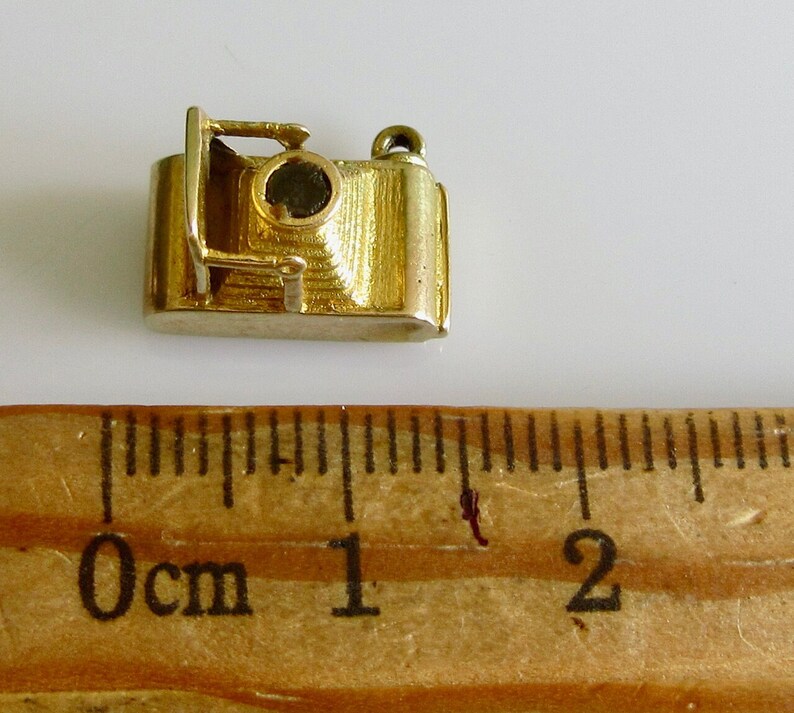 May include: A gold-colored camera charm with a black lens. The charm is about 1 inch long and is sitting on a wooden ruler.