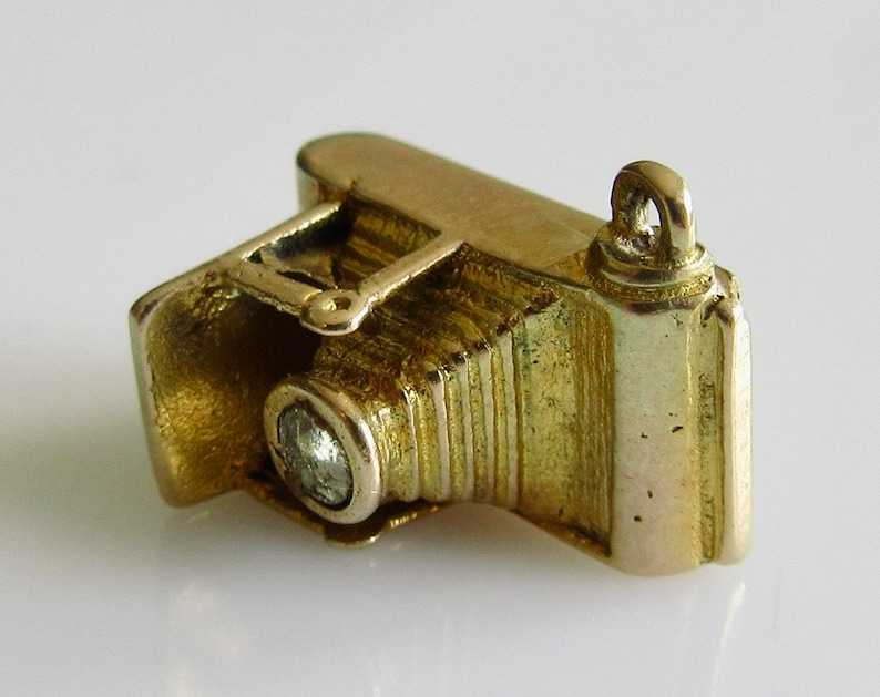 May include: A gold charm in the shape of an old-fashioned camera with a single diamond-shaped stone in the lens.