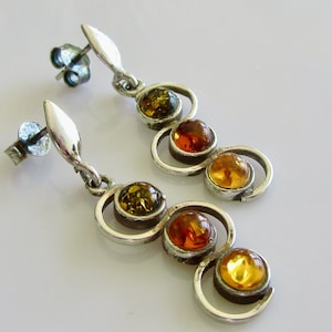 Puede incluir: Pendientes colgantes con metal plateado y piedras de ámbar. Cada pendiente presenta tres piedras de ámbar en tonos amarillo, naranja y verde, engastadas en un diseño en espiral. Cierre de poste. Aproximadamente 5 cm de largo.