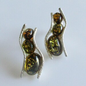 May include: A pair of silver earrings with three amber-coloured stones each. The stones are oval and round, with dark brown and olive green hues. The earrings have a curved, openwork design.