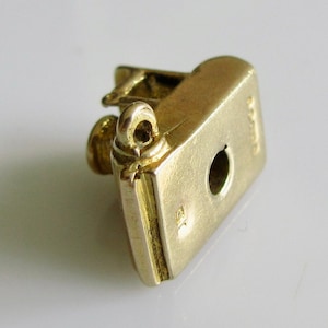 May include: Gold charm in the shape of a book with a loop on top. The charm has a hole in the bottom for attaching to a bracelet or necklace. The charm is marked with the number "925" on the side.