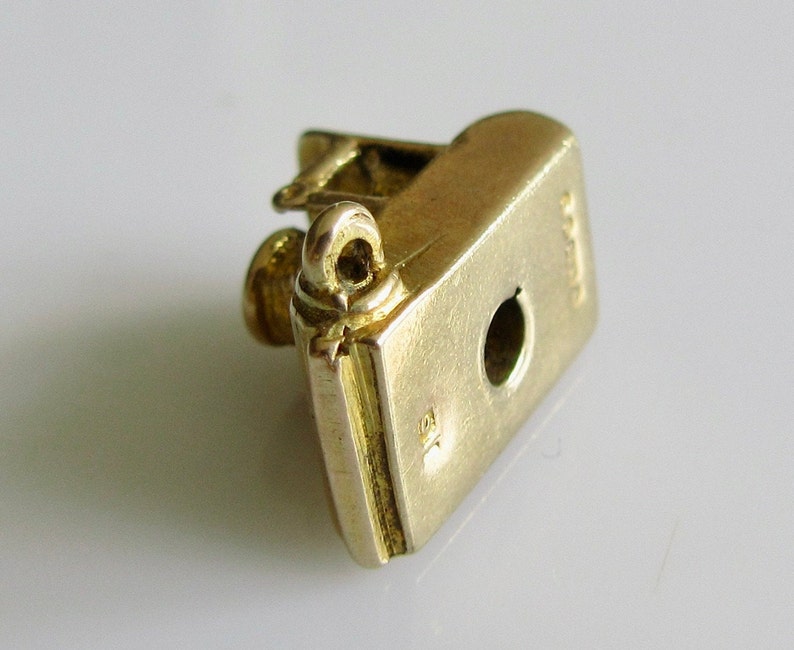May include: Gold charm in the shape of a book with a loop on top. The charm has a hole in the bottom for attaching to a bracelet or necklace. The charm is marked with the number "925" on the side.
