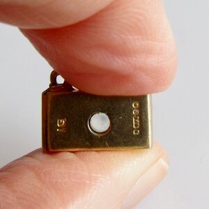 May include: A gold charm in the shape of a small book with a round, clear stone in the center. The charm has the inscription "925" and "IG" on the back.