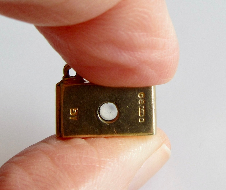 May include: A gold charm in the shape of a small book with a round, clear stone in the center. The charm has the inscription "925" and "IG" on the back.
