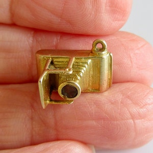 May include: A gold-colored charm in the shape of an old-fashioned camera. The camera has a lens and a bellows.