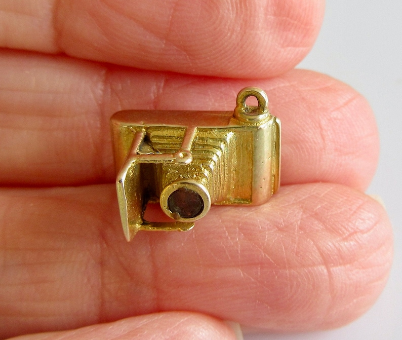 May include: A gold-colored charm in the shape of an old-fashioned camera. The camera has a lens and a bellows.