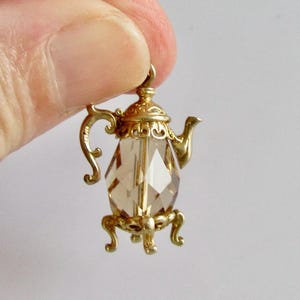 May include: A gold-coloured teapot charm with a faceted, amber-coloured crystal body. The teapot has ornate details, including a curved handle, a spout, and decorative feet. The charm is held by a finger against a white background.