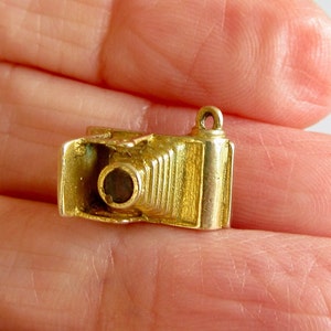 May include: A gold-colored charm in the shape of an old-fashioned camera. The camera has a round lens and a bellows design.
