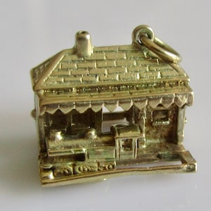 May include: A gold-colored charm depicting a train station with a train pulling into the station. The charm is detailed with a brick roof, a small window, and a train with wheels.