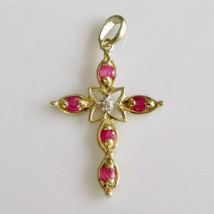 May include: A gold cross pendant with a floral design. The cross features oval, bright red gemstones and a small, clear gemstone in the centre. The pendant has a loop at the top for a chain.