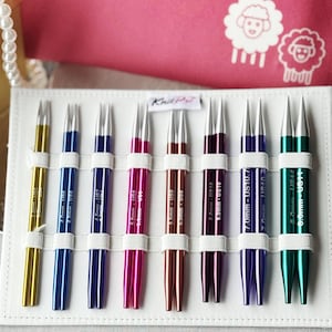 Peut inclure: Un ensemble d'aiguilles à tricoter Knit Pro dans un étui blanc. Les aiguilles sont de différentes couleurs, dont l'or, le bleu, le rose et le vert. Chaque aiguille est étiquetée avec sa taille en millimètres et en tailles US. Un étui rose avec des motifs de moutons est en arrière-plan.