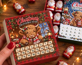 May include: A square Christmas countdown calendar box featuring a Highland cow illustration, red plaid borders, and numbered squares from 1 to 24. The text reads Christmas Countdown To Go. Small foil-wrapped chocolate Santa figures are scattered nearby.