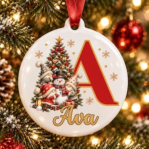 Può includere: Una decorazione natalizia rotonda in ceramica bianca con una grande lettera A rossa, il nome Ava in corsivo dorato e un'illustrazione festiva di un albero di Natale decorato, un pupazzo di neve e due gnomi, appesa con un nastro rosso.
