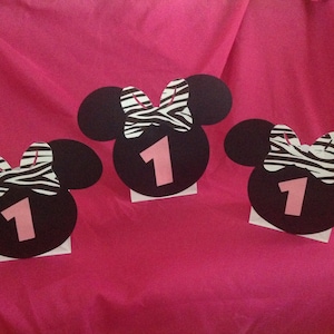May include: Three black Minnie Mouse-shaped decorations with pink number 1 cutouts. Each has a zebra print bow and is mounted on a white base. The decorations are arranged on a bright pink background, suitable for a first birthday party.