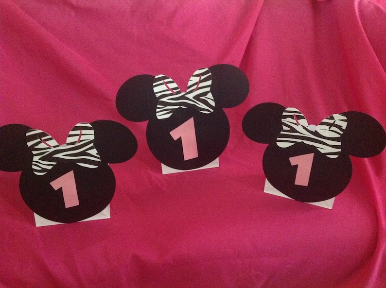 May include: Three black Minnie Mouse-shaped decorations with pink number 1 cutouts. Each has a zebra print bow and is mounted on a white base. The decorations are arranged on a bright pink background, suitable for a first birthday party.
