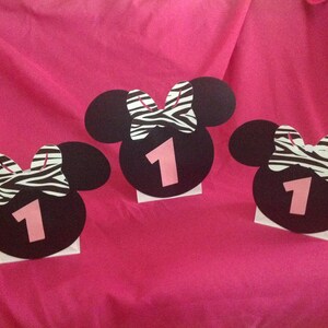 May include: Three black Minnie Mouse head cutouts with zebra print bows and the number 1 in pink. The cutouts are on white bases, set against a bright pink background, likely for a first birthday party.