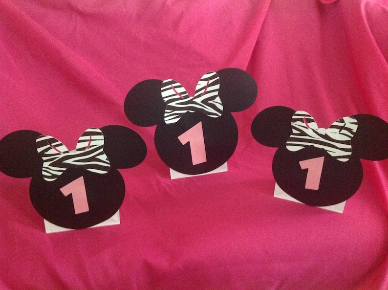 May include: Three black Minnie Mouse head cutouts with zebra print bows and the number 1 in pink. The cutouts are on white bases, set against a bright pink background, likely for a first birthday party.