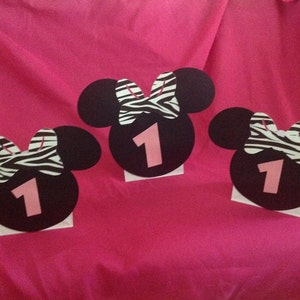 May include: Three black Minnie Mouse-shaped decorations with a pink number 1 and a zebra-striped bow. The decorations are on a white base and set against a pink background, suitable for a first birthday party.