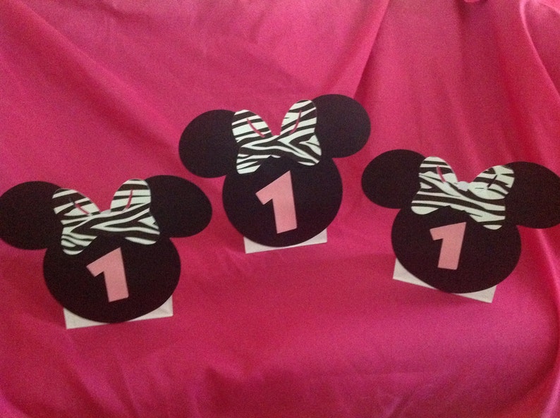 May include: Three black Minnie Mouse-shaped decorations with a pink number 1 and a zebra-striped bow. The decorations are on a white base and set against a pink background, suitable for a first birthday party.