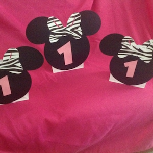 May include: Three black Minnie Mouse-shaped decorations with a zebra print bow and the number 1 in pink. Each decoration is mounted on a small white square, set against a bright pink background.