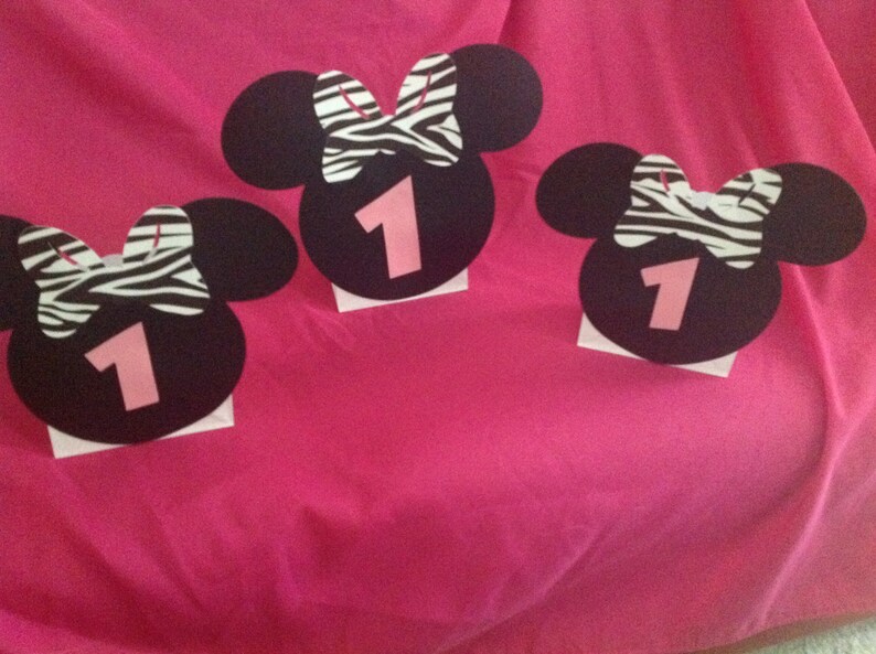 May include: Three black Minnie Mouse-shaped decorations with a zebra print bow and the number 1 in pink. Each decoration is mounted on a small white square, set against a bright pink background.