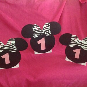 May include: Three black Mickey Mouse head cutouts with a zebra print bow and the number 1 in pink. The cutouts are on a pink background, likely for a birthday party or celebration.