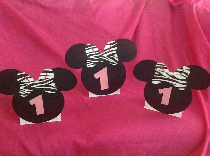 May include: Three black Mickey Mouse head cutouts with a zebra print bow and the number 1 in pink. The cutouts are on a pink background, likely for a birthday party or celebration.