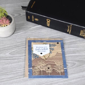 May include: A greeting card with a landscape design, featuring trees and mountains, framed in blue and brown. The card includes the text "Wishing you God's richest blessings." A black Bible and a small succulent plant in a white pot are also visible.