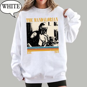 Może przedstawiać: Biała bluza z nadrukiem The Mandalorian. Projekt zawiera napis "THE MANDALORIAN" w stylu vintage, czarno-biały obraz postaci oraz napis "EST. 2019" z poziomymi paskami.