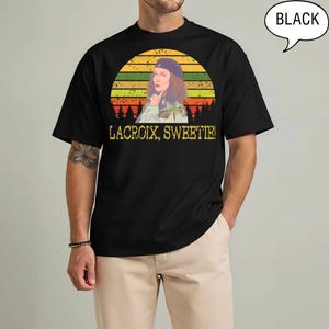 May include: A black short-sleeve t-shirt featuring a graphic of a person wearing a beret in front of a sunset and forest silhouette. Below the graphic, the text reads LACROIX, SWEETIE! in yellow letters. The shirt has a crew neckline.