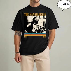 Może przedstawiać: Czarny t-shirt z grafiką w stylu retro The Mandalorian. Projekt zawiera złoty napis "THE MANDALORIAN", wizerunek postaci oraz złoty napis "EST. 2019". Koszulka ma okrągły dekolt.