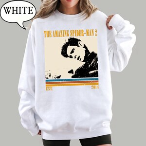 May include: White sweatshirt with a graphic from "The Amazing Spider-Man 2." The design features a black and beige image of a person, with text and stripes in orange, blue, and brown. The text "EST. 2014" is also included.