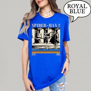 May include: Royal blue t-shirt featuring a graphic from the movie Spider-Man 2. The design includes the movie title, a building scene, and the year 2004. The shirt is a solid royal blue color.
