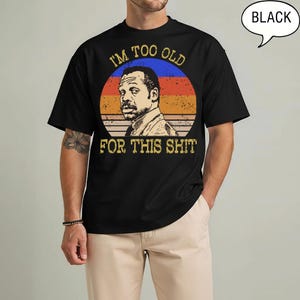 May include: A black t-shirt featuring a circular graphic with a distressed sunset background in blue, orange, and red. The graphic depicts a man with a mustache and the text I'M TOO OLD FOR THIS SHIT in a distressed yellow font above and below.