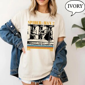 May include: Ivory-colored t-shirt featuring a vintage-style graphic from Spider-Man 2. The design includes the film's title, a building illustration, and the text "EST. 2004" in a retro font. The shirt is worn by a person with blonde hair.