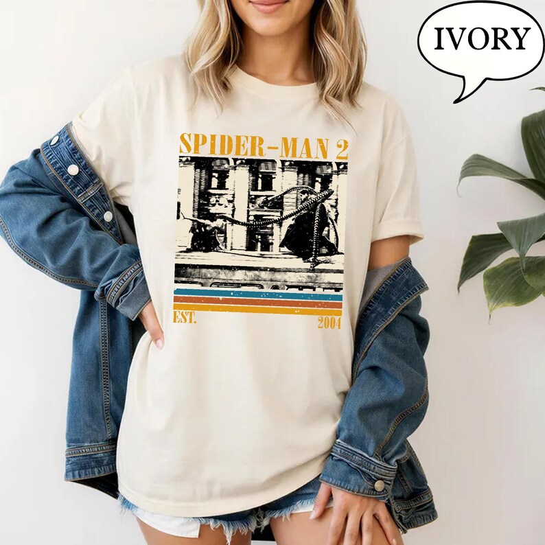 May include: Ivory-colored t-shirt featuring a vintage-style graphic from Spider-Man 2. The design includes the film's title, a building illustration, and the text "EST. 2004" in a retro font. The shirt is worn by a person with blonde hair.
