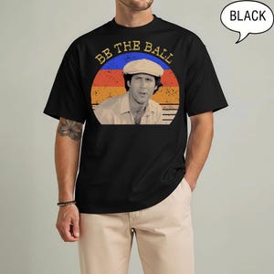 May include: A black short-sleeved t-shirt featuring a circular graphic with a sunset-colored background of orange, red, and blue stripes. The graphic depicts a person wearing a light-colored flat cap and includes the text BE THE BALL.