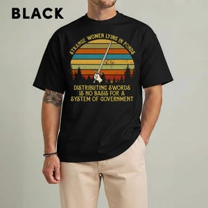 May include: Black t-shirt with a graphic featuring a sword and the text "STRANGE WOMEN LYING IN PONDS DISTRIBUTING SWORDS IS NO BASIS FOR A SYSTEM OF GOVERNMENT" in a retro sunset design. The word "BLACK" is printed at the top.