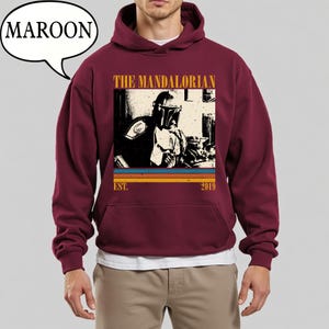 Może przedstawiać: Bordowa bluza z kapturem z grafiką The Mandalorian. Projekt zawiera wizerunek postaci, napis "THE MANDALORIAN" oraz rok "2019". Bluza posiada przednią kieszeń i kaptur ze sznurkiem.