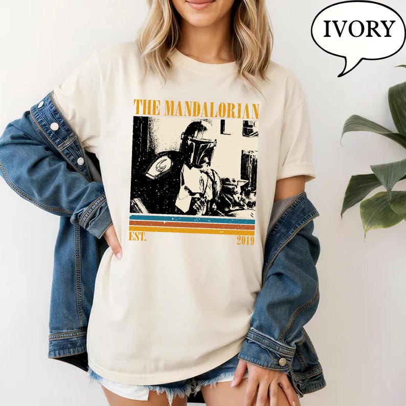 Może przedstawiać: Kremowa koszulka z czarno-białą grafiką The Mandalorian. Projekt zawiera napis "THE MANDALORIAN" nad obrazem i "EST. 2019" poniżej, z poziomymi paskami w kolorze pomarańczowym, niebieskim i brązowym.
