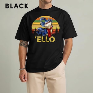 May include: Black t-shirt featuring a colorful graphic of a blue and white worm-like creature wearing a red scarf, set against a sunset design. The word "'ELLO" is printed in yellow below the creature. The word "BLACK" is printed in white at the top.