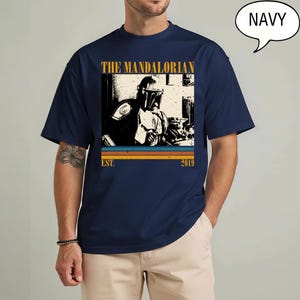 Może przedstawiać: Granatowa koszulka z nadrukiem The Mandalorian. Projekt zawiera złoty napis "THE MANDALORIAN" z napisem "EST. 2019" poniżej. Obraz ma wygląd vintage z poziomymi paskami w kolorze pomarańczowym, ż&oacute;łtym i niebieskim.