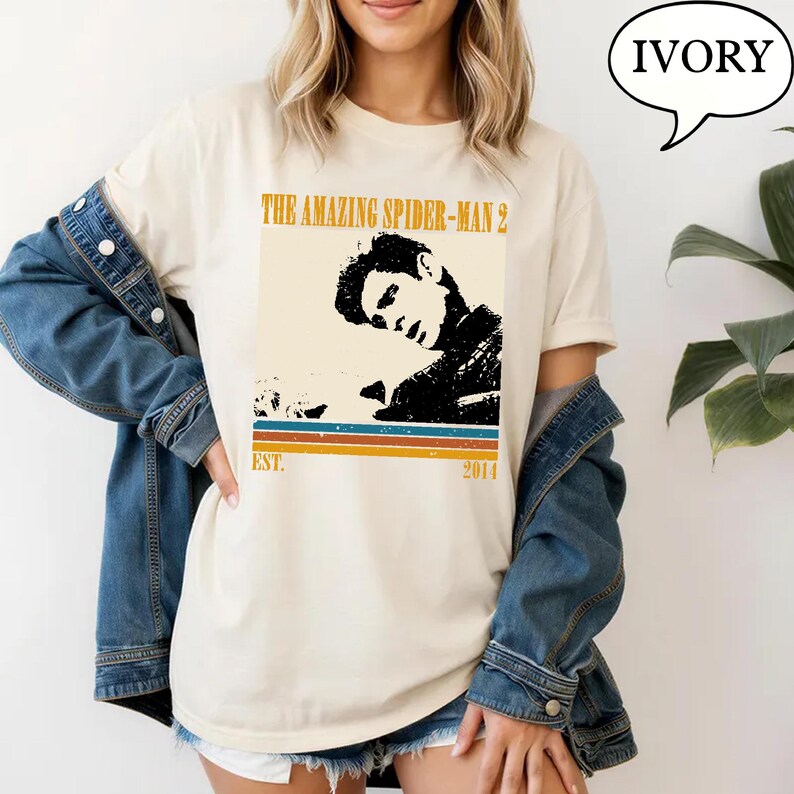 May include: Ivory t-shirt with a graphic from "The Amazing Spider-Man 2." The design features a black and white image of a person, with text above and below the image. The text reads "THE AMAZING SPIDER-MAN 2" and "EST. 2014."