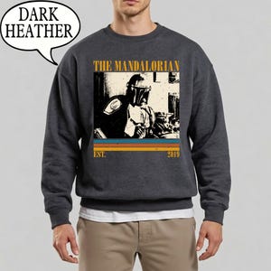 Może przedstawiać: Ciemnoszara bluza z nadrukiem The Mandalorian. Projekt zawiera napis "THE MANDALORIAN" nad wizerunkiem postaci, z napisem "EST. 2019" poniżej. Bluza ma okrągły dekolt.
