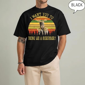 May include: A black short-sleeve t-shirt featuring a graphic of a knight in a helmet with antlers. The design includes a sunset background, a forest silhouette, and the text: I WANT YOU TO BRING ME A SHRUBBERY. A speech bubble says BLACK.