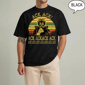 May include: A black short-sleeve t-shirt featuring a graphic of a Martian character with a large brain. The design includes a sunset background with horizontal stripes and the repeated yellow text "ACK ACK" and "ACK ACKACK ACK".