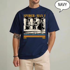 May include: Navy blue t-shirt featuring a graphic from Spider-Man 2. The design includes the text "SPIDER-MAN 2" in gold, a black and white image, and the text "EST. 2004" in a retro style.
