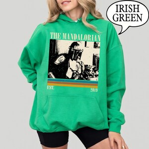 Może przedstawiać: Zielona bluza z kapturem w kolorze irlandzkiej zieleni z grafiką The Mandalorian. Grafika jest czarno-biała, z napisem "THE MANDALORIAN" nad obrazem i "EST. 2019" poniżej. Bluza ma kaptur ze sznurkiem i długie rękawy.