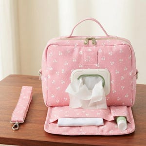May include: A pink fabric travel case with a white floral pattern, featuring a top handle, a zippered main compartment, and a front-facing dispenser for wet wipes. The case includes a detachable wrist strap and an open front pocket with a white cloth.